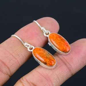 Könnte beinhalten: Ein Paar silberne Ohrhänger mit ovalen, orangefarbenen Edelsteinen. Die Edelsteine haben ein gesprenkeltes Aussehen. Die Ohrringe werden von den Fingern einer Person vor einem dunklen Hintergrund gehalten. Schmuck.