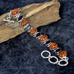 May include: A silver bracelet with oval amber and dark red gemstone details. The bracelet has a toggle clasp and is displayed on wood against a dark blue backdrop. The amber stones have a warm, golden-orange hue.