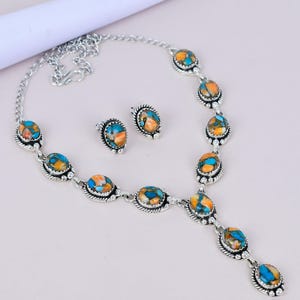 May include: A silver necklace and earring set. The jewellery features oval stones in blue, orange, and brown. The necklace has a pendant design, and the earrings are studs. The set is displayed on a light background. The necklace and earrings are made of silver.