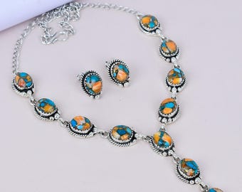May include: A silver-toned necklace and matching earrings set. The jewelry features oval-shaped stones with a mix of blue, orange, and brown hues. The necklace has a pendant design, and the earrings are studs. The set is displayed on a light background.