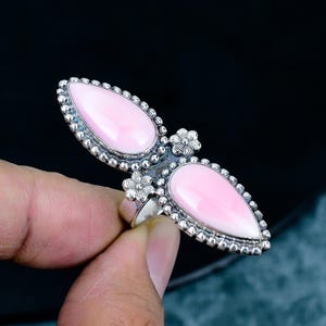 May include: A silver ring featuring two elongated, pear-shaped pink stones set in a beaded metal frame. The centre of the ring is decorated with two small, detailed metal flowers positioned between the two smooth, light pink cabochon stones.