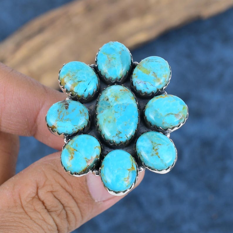 Puede incluir: Anillo de plata con piedras turquesas. El anillo presenta nueve piedras turquesas ovaladas y redondas, dispuestas en forma de flor. Las piedras turquesas son de color azul-verdoso con vetas marrones.