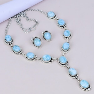 May include: A silver necklace and matching stud earrings set. The jewellery features oval, light blue gemstones set in ornate silver settings. The necklace has a pendant design with a dangling gemstone. The earrings are small and round.
