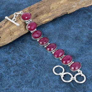 Op de afbeelding: Een zilveren armband met een ketting en een sluiting, met zeven ovale, donkerroze edelstenen.