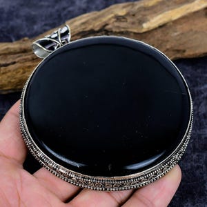 Puede incluir: Un colgante grande y redondo de ónix negro con un borde decorativo de color plateado. El colgante tiene un lazo en la parte superior para una cadena. El colgante se sostiene en una mano sobre un fondo oscuro.