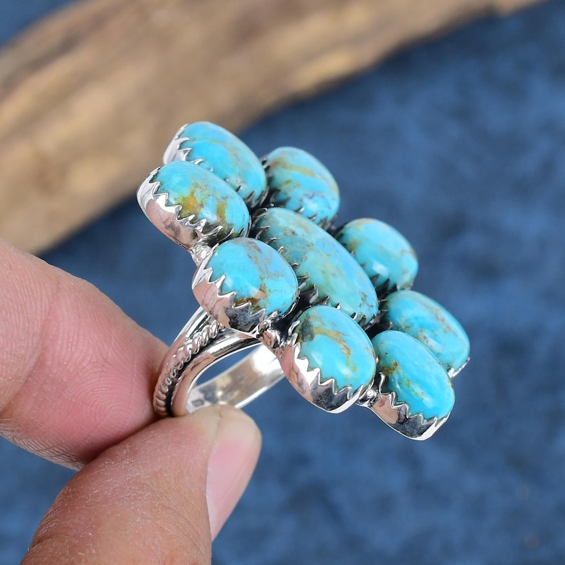 Puede incluir: Un anillo de plata con un grupo de piedras turquesas. El anillo tiene una banda detallada y una disposici&oacute;n floral de piedras turquesas ovaladas de color azul claro con vetas marrones. El anillo es sostenido por una mano sobre un fondo azul.
