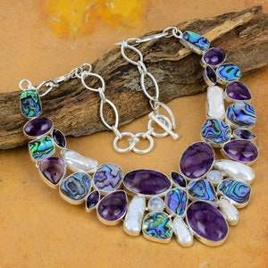May include: A silver necklace with a statement design featuring a mix of purple amethyst, iridescent abalone shell, and white freshwater pearls. The necklace has a lobster clasp closure and a silver chain.