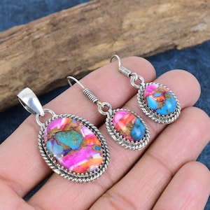 May include: A set of silver earrings and a pendant featuring a colourful oval-shaped stone with pink, blue, and orange hues. The stone is set in a silver bezel with a decorative border.