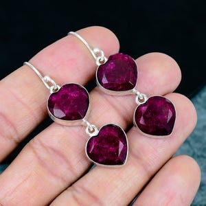 May include: A pair of drop earrings featuring faceted, heart-shaped deep magenta stones set in silver-toned metal. Each earring consists of two heart-shaped stones linked vertically, suspended from a simple silver-toned hook wire finding.