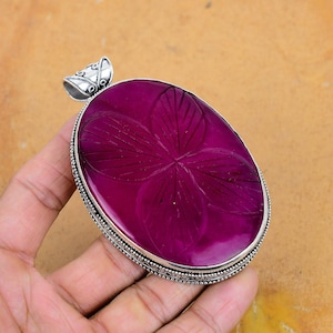 以下が含まれることがあります： 深いマゼンタ色の宝石が付いた、大きな楕円形のペンダント。石には彫刻された花の模様があり、装飾的なベイルが付いた銀色の金属フレームにセットされています。ペンダントはニュートラルな背景に対して保持されています。