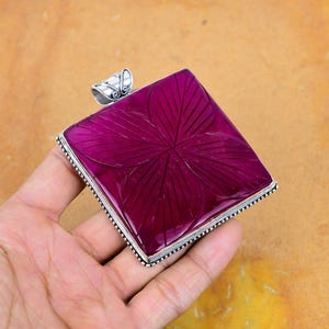 Peut inclure: Un pendentif carré magenta avec un motif floral sculpté. Le pendentif est serti dans un cadre en métal argenté avec une bélière décorative. Le pendentif est tenu dans une main sur un fond chaud et neutre.