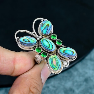 May include: A silver-toned ring featuring a butterfly design with oval-shaped abalone shell stones in shades of blue and green. Small, round, green stones are interspersed among the larger stones. The ring has a textured band.