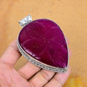 Könnte beinhalten: Ein großer, tropfenförmiger Anhänger mit einem tiefmagentafarbenen Edelstein. Der Stein weist ein geätztes Blumenmuster auf und ist in einen silberfarbenen Metallrahmen gefasst. Der Anhänger hat eine dekorative Öse.