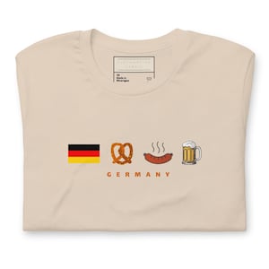 Op de afbeelding: Een beige T-shirt met een afbeelding van een Duitse vlag, een pretzel, een worst en een bierglas. De tekst "GERMANY" staat onder de afbeelding.