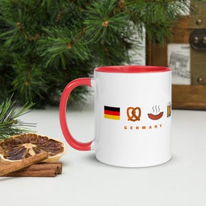 Op de afbeelding: Witte keramische mok met een rode binnenkant en handvat. De mok toont de Duitse vlag, een pretzel, een worst en een bierpul. Het woord "GERMANY" staat eronder gedrukt.