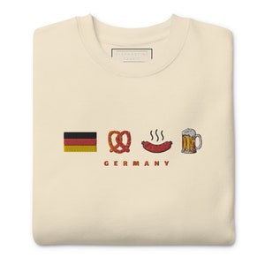 Op de afbeelding: Crèmekleurige sweatshirt met geborduurde Duitse vlag, pretzel, worst en bierpul. Het woord "GERMANY" is eronder geborduurd. Het sweatshirt heeft een label met de tekst "ELEPHANTINE FABRIC".