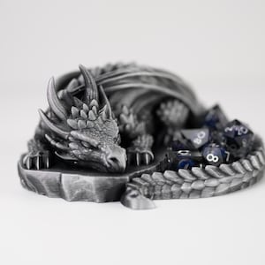 A detailed side-profile view of the "Nap Twenty" sleeping dragon dice holder. The 3D-printed dragon features a metallic silver finish, showcasing intricate scales, curved horns, and a peaceful expression. A set of shimmering purple and black polyhedral dice is tucked into the center of its coiled body. The dragon is resting on a light gray surface, emphasizing its role as a decorative and functional tabletop gaming accessory.