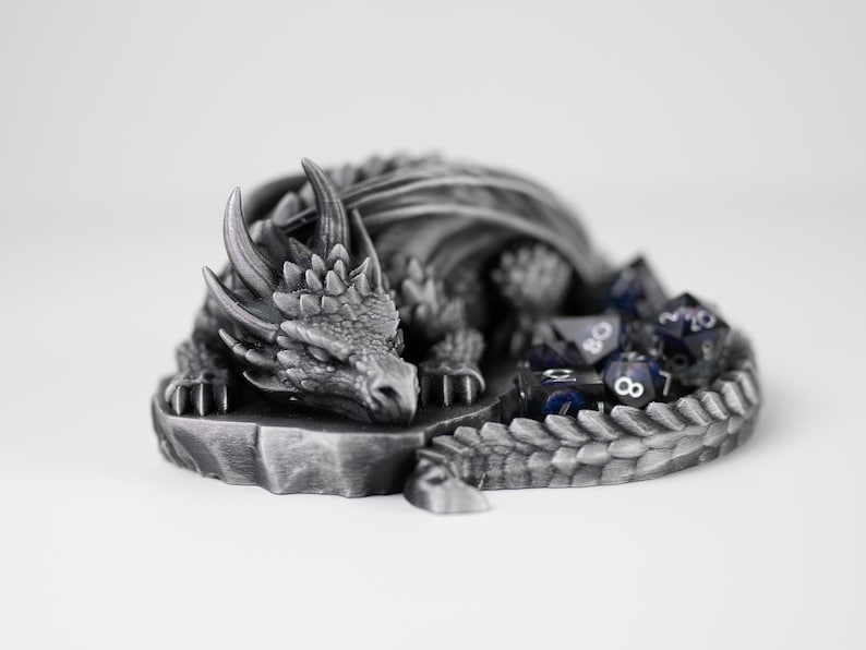 A detailed side-profile view of the "Nap Twenty" sleeping dragon dice holder. The 3D-printed dragon features a metallic silver finish, showcasing intricate scales, curved horns, and a peaceful expression. A set of shimmering purple and black polyhedral dice is tucked into the center of its coiled body. The dragon is resting on a light gray surface, emphasizing its role as a decorative and functional tabletop gaming accessory.