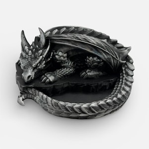 A 3D-printed "Nap Twenty" sleeping dragon dice holder in an antique silver finish. The dragon is curled in a circular "O" shape, creating a central basin to hold gaming dice. The high-detail print shows off individual scales, spinal ridges, and a peaceful facial expression. The model is resting on a plain white background, emphasizing the metallic sheen and weathered texture of the silver filament.