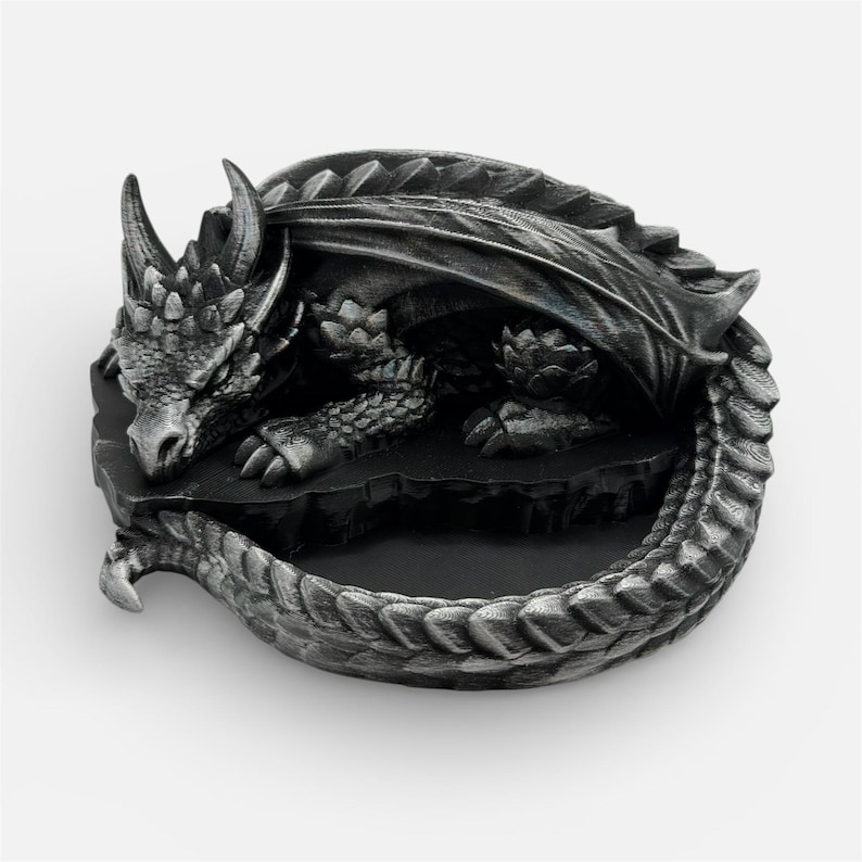 A 3D-printed "Nap Twenty" sleeping dragon dice holder in an antique silver finish. The dragon is curled in a circular "O" shape, creating a central basin to hold gaming dice. The high-detail print shows off individual scales, spinal ridges, and a peaceful facial expression. The model is resting on a plain white background, emphasizing the metallic sheen and weathered texture of the silver filament.