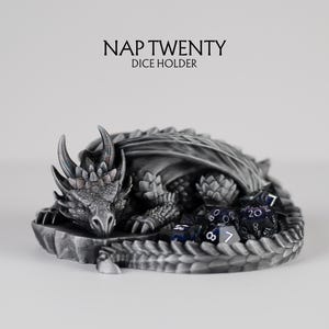 A 3D-printed "Nap Twenty" dice holder featuring a highly detailed sleeping dragon curled in a circle. The dragon has a metallic silver and charcoal finish with realistic scales and horns. A set of purple and black translucent polyhedral dice is nestled within the curve of the dragon's tail. The text above the dragon reads "NAP TWENTY DICE HOLDER" against a clean, light grey background.