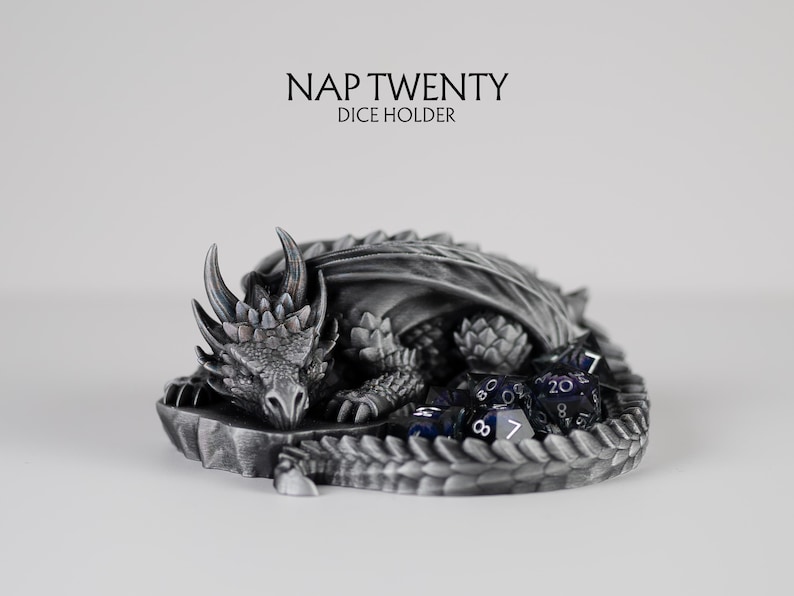 A 3D-printed "Nap Twenty" dice holder featuring a highly detailed sleeping dragon curled in a circle. The dragon has a metallic silver and charcoal finish with realistic scales and horns. A set of purple and black translucent polyhedral dice is nestled within the curve of the dragon's tail. The text above the dragon reads "NAP TWENTY DICE HOLDER" against a clean, light grey background.