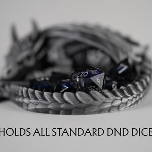 A close-up, shallow depth-of-field shot focusing on the tail of the metallic silver 3D-printed dragon. The text "HOLDS ALL STANDARD DND DICE" is displayed at the bottom. Inside the dragon's coiled tail, a translucent purple d20 die is in sharp focus, while the rest of the sleeping dragon and other dice in the set are softly blurred in the background.