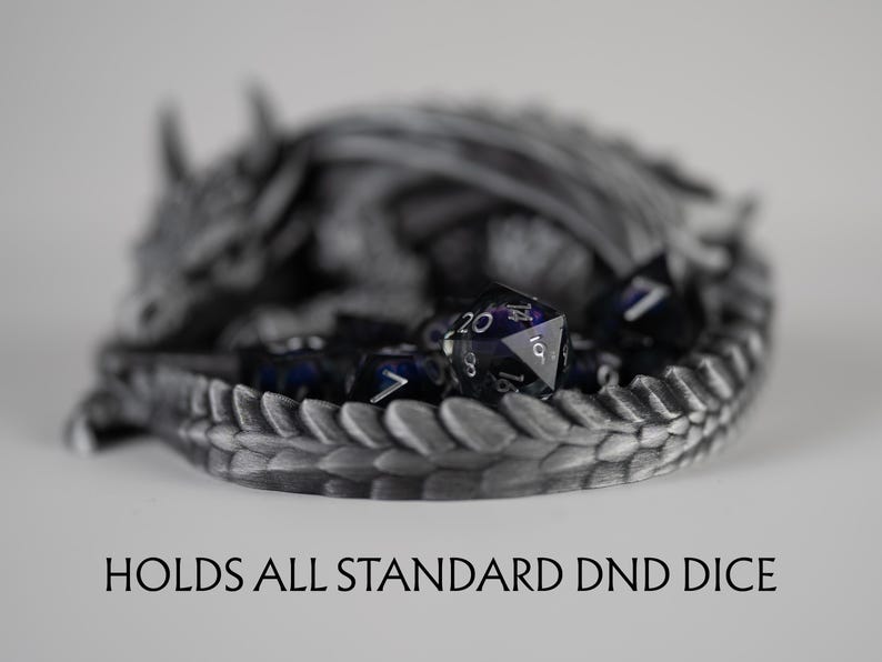 A close-up, shallow depth-of-field shot focusing on the tail of the metallic silver 3D-printed dragon. The text "HOLDS ALL STANDARD DND DICE" is displayed at the bottom. Inside the dragon's coiled tail, a translucent purple d20 die is in sharp focus, while the rest of the sleeping dragon and other dice in the set are softly blurred in the background.