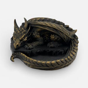 A top-down, angled view of the "Nap Twenty" 3D-printed dragon dice holder in an antique gold finish. The sleeping dragon is coiled into a circle, with its head resting near its tail, creating a central basin for dice. The metallic gold filament highlights the intricate scale textures and spinal ridges, giving it a weathered, treasure-like appearance against a plain white background.