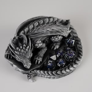 An elevated, top-down view of the "Nap Twenty" 3D-printed dragon dice holder. The sleeping dragon is curled in a tight circle, showing off intricate scale details and spinal ridges in a metallic silver finish. A full set of dark purple galaxy-themed polyhedral dice is held securely within the center of the dragon's coiled body and tail.