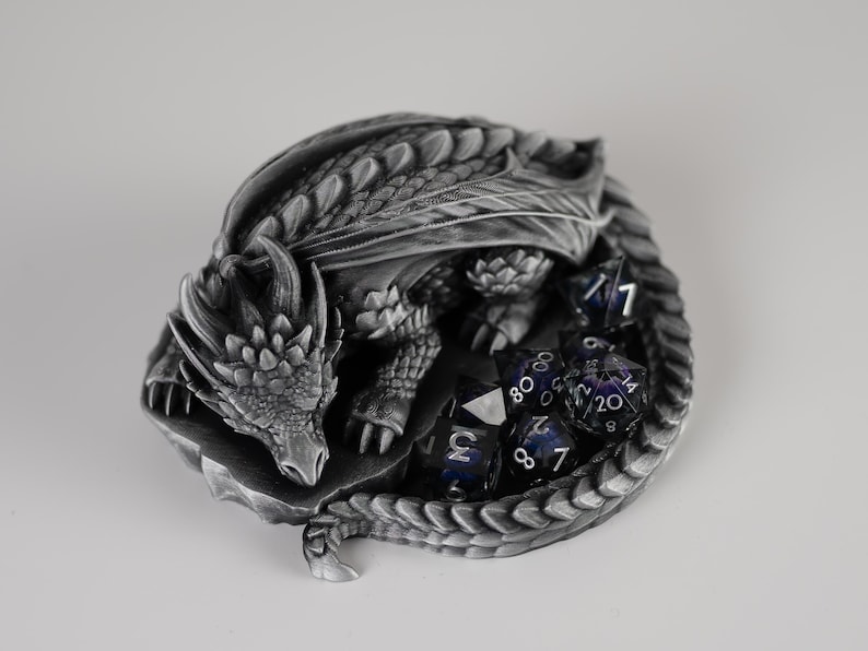 An elevated, top-down view of the "Nap Twenty" 3D-printed dragon dice holder. The sleeping dragon is curled in a tight circle, showing off intricate scale details and spinal ridges in a metallic silver finish. A full set of dark purple galaxy-themed polyhedral dice is held securely within the center of the dragon's coiled body and tail.