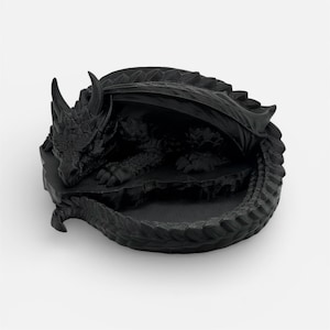 A top-down, angled view of the "Nap Twenty" 3D-printed dragon dice holder in a matte black finish. The sleeping dragon is curled in a circle, highlighting the sharp, clean lines of its scales and horns. The central hollow for holding dice is empty, showcasing the interior detail of the 3D-printed model against a neutral white background.