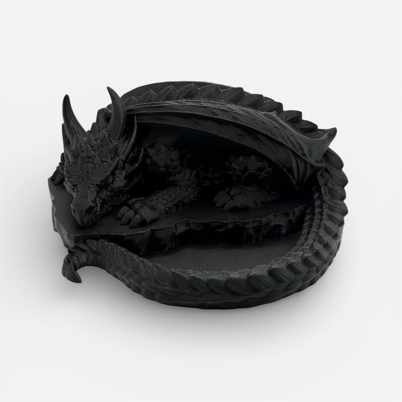 A top-down, angled view of the "Nap Twenty" 3D-printed dragon dice holder in a matte black finish. The sleeping dragon is curled in a circle, highlighting the sharp, clean lines of its scales and horns. The central hollow for holding dice is empty, showcasing the interior detail of the 3D-printed model against a neutral white background.