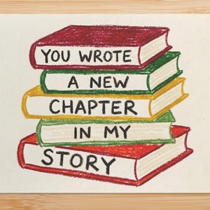 May include: A greeting card featuring a stack of illustrated books. Each book is a different color, with the text "YOU WROTE A NEW CHAPTER IN MY STORY" written on the spines. The card is on a wooden surface.