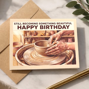 May include: A birthday card with the text "STILL BECOMING SOMETHING BEAUTIFUL HAPPY BIRTHDAY" features an illustration of hands shaping clay on a pottery wheel. The card is accompanied by a brown envelope and a gold pen.