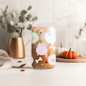 May include: A clear glass tumbler with a Halloween-themed design. The tumbler features ghosts, cauldrons, bones, and skulls in shades of pink, green, and black. The tumbler is filled with a brown liquid.