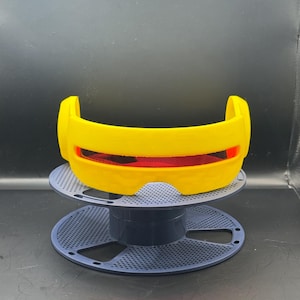 May include: A yellow, 3D-printed visor with a red lens, resting on a blue spool-shaped stand. The visor has a curved design and is likely a cosplay or costume accessory. The stand has a perforated pattern.