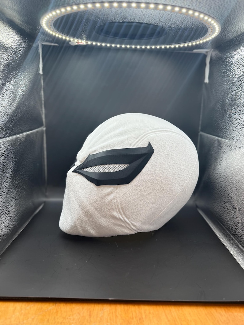 以下が含まれることがあります： 黒いアイマスクが付いた白いヘルメット。ヘルメットはテクスチャ加工された表面を持ち、リングライトで照らされています。アイマスクはメッシュデザインです。