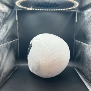 以下が含まれることがあります： 白の野球帽、つばはカーブしており、黒いパッチが付いています。帽子は質感のある素材でできており、リングライトを備えたスタジオボックス内の黒い背景に配置されています。