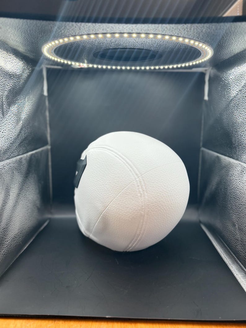 以下が含まれることがあります： 白の野球帽、つばはカーブしており、黒いパッチが付いています。帽子は質感のある素材でできており、リングライトを備えたスタジオボックス内の黒い背景に配置されています。