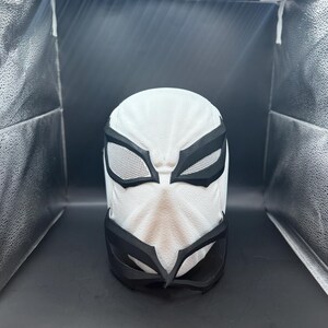 以下が含まれることがあります： 白と黒のスーパーヒーローマスク。表面はテクスチャ加工が施されています。マスクには黒い目と口のディテールがあり、目の開口部を覆うメッシュのような素材があります。マスクは頭全体を覆うように設計されています。