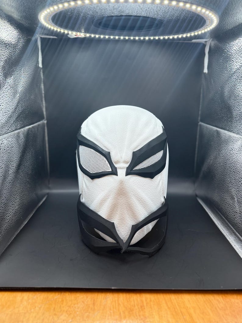 以下が含まれることがあります： 白と黒のスーパーヒーローマスク。表面はテクスチャ加工が施されています。マスクには黒い目と口のディテールがあり、目の開口部を覆うメッシュのような素材があります。マスクは頭全体を覆うように設計されています。