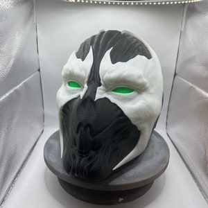 May include: A black and white mask with green eyes, reminiscent of a comic book character. The mask is displayed on a gray stand, showcasing its intricate design and details. The mask's design features a combination of sharp angles and smooth curves.