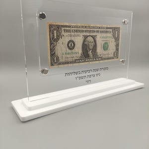 May include: A clear acrylic frame with a one dollar bill inside. The bill is a Federal Reserve Note from the United States of America. The bill features a portrait of George Washington and the text "ONE DOLLAR". The frame has a white base with the text "מזכרת שנת רביעית בשליחות ניס צרפת תשס"ו דוד" in Hebrew.