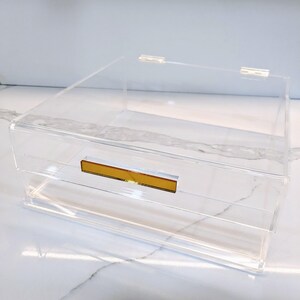 May include: A clear acrylic storage box with a gold drawer pull. The box has a lid and two drawers.