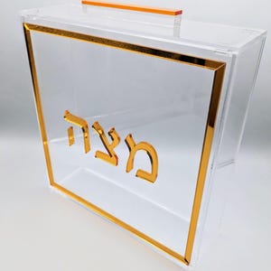 Peut inclure: Une boîte en acrylique transparente avec une bordure dorée et une inscription dorée qui dit "מצה" en hébreu. La boîte a une petite poignée orange sur le dessus.