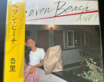 Può includere: Una copertina di un disco in vinile con sfondo giallo e una foto di una donna che tiene una borsa di carta marrone. Il testo sulla copertina recita "Heaven Beach ANRI" in lettere nere e rosse. Il testo "ヘブン・ビーチ 杏里" è anche sulla copertina in giapponese.