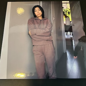 May include: A person wearing a mauve-colored hoodie and matching sweatpants, leaning against a gray wall. The outfit appears to be made of a soft material. The person has short, dark hair.