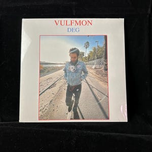 May include: A sealed vinyl record album featuring the text "VULFMON DEG" in red and blue. The album cover shows a person walking down a road with a small dog in their jacket pocket. The background includes trees and a fence.