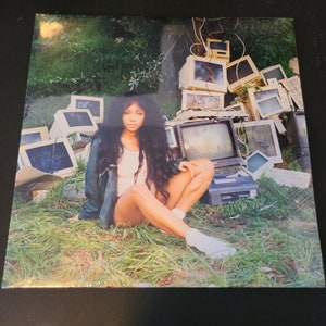 May include: A woman with long dark hair sits in a field of green grass. She is wearing a white tank top, a dark green jacket, and white socks. Behind her is a pile of old computer monitors and other electronics.